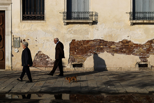 Two elderly people are walking along a street, slightly separated from each other.